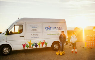 Das Spielmobil auf einer Straße vor dem Sonnenuntergang. Zwei Kinder spielen davor mit Spielsachen.