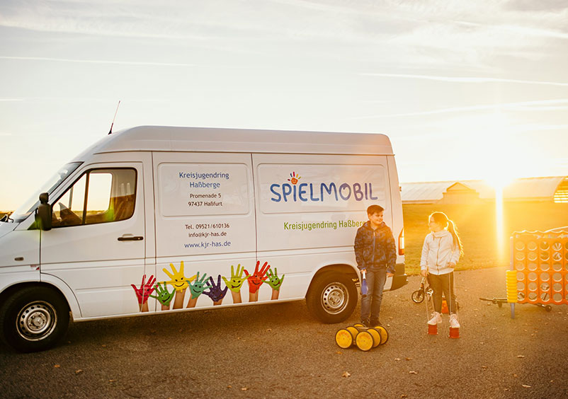 Das Spielmobil auf einer Straße vor dem Sonnenuntergang. Zwei Kinder spielen davor mit Spielsachen.