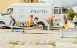 Zwei Kinder rutschen auf einer Rollenrutsche im Fokus der Kamera. Das Spielmobil im Hintergrund ist verschwommen, dadurch wirken die Kinder schnell.
