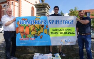 Hofheims Bürgermeister Wolfgang Borst, Vorstandsmitglied Benjamin Gerschütz und Vorsitzender Thomas Wagenhäuser halten gemeinsam am Hofheimer Marktplatz ein Bauzaunbanner der Haßberger Spieletage hoch