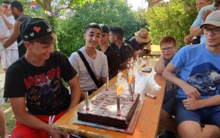 Ein Kuchen mit Kerzen als Geburtstagsüberraschung für einen Jugendlichen, der gemeinsam mit anderen auf einer Biertischgarnitur sitzt.
