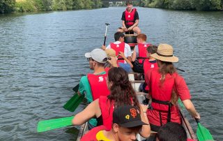 Die Teilnehmenden der Jugendbegegnung sitzen in Schwimmwesten auf einem Boot und rudern. Der Steuermann sitzt vorne erhöht
