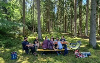 Neun Personen sitzen an und um eine Biertischgarnitur im Wald und essen