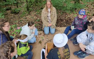Spielerisches kennenlernen des Elements Erde, bei dem einige Kinder im Wald auf einem Karton sitzen.