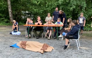 Einige Kinder spielen ein Quiz, während eine Person vor ihnen als Rätsel am Boden liegt