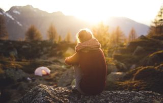 Eine Person sitzt auf einem Stein und betrachtet die hinter Bergen untergehende Sonne