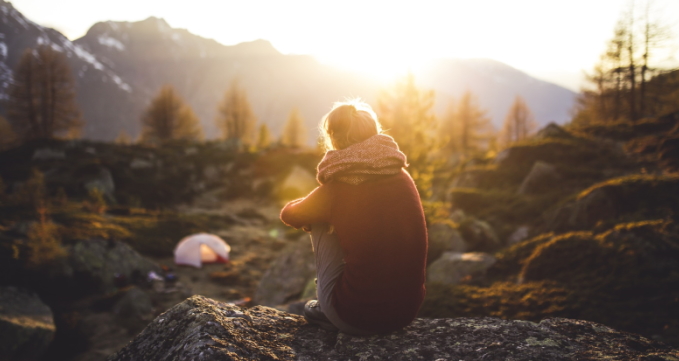 Eine Person sitzt auf einem Stein und betrachtet die hinter Bergen untergehende Sonne