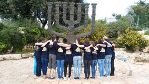 Eine Gruppe der Jugendbegegnung mit Israel steht mit dem Rücken zur Kamera Arm in Arm vor einem großen Chanukka Leuchter.