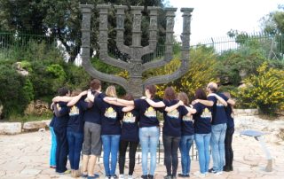Eine Gruppe der Jugendbegegnung mit Israel steht mit dem Rücken zur Kamera Arm in Arm vor einem großen Chanukka Leuchter.