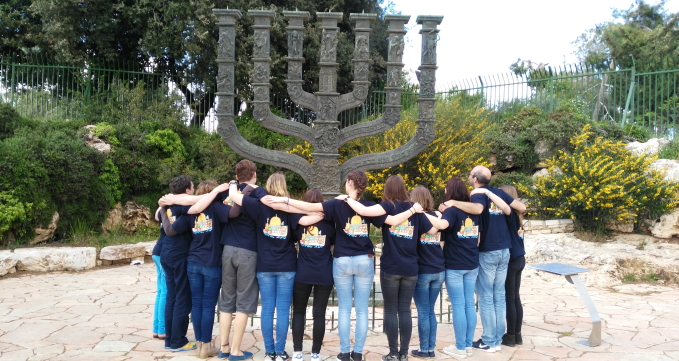 Eine Gruppe der Jugendbegegnung mit Israel steht mit dem Rücken zur Kamera Arm in Arm vor einem großen Chanukka Leuchter.