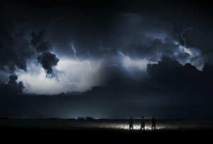 Drei Personen stehen in einem Feld bei Nacht und am Himmel sind Blitze und Wolken zu sehen