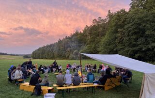 Jugendliche sitzen am Lagerfeuer beim Zeltlager in Reutersbrunn vor einem lila Himmel