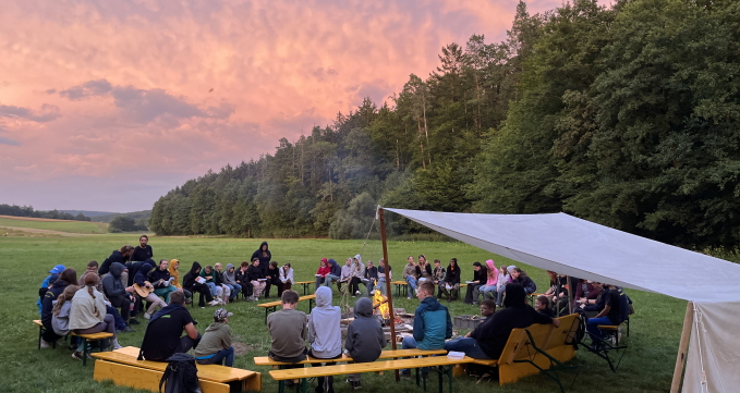 Jugendliche sitzen am Lagerfeuer beim Zeltlager in Reutersbrunn vor einem lila Himmel