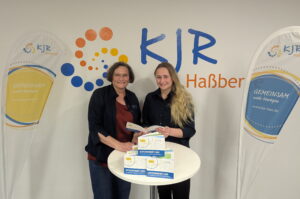 Kreisjugendring Vorsitzende Susanne Makowski präsentiert gemeinsam mit Vorstandsmitglied Anna-Maria Gebhardt das Juleica-Gutscheinheft 2025 vor dem Logo des Kreisjugendring Haßberge.