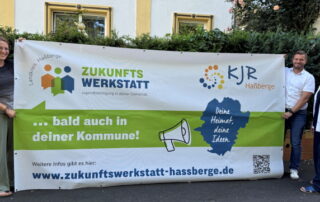 Eva Pfeil (KJR-Geschäftsführerin, Kreisjugendpflegerin), Thomas Wagenhäuser (stellvertretender KJR-Vorsitzender) und Susanne Makowski (KJR-Vorsitzende) halten das Bauzaunbanner für die 2025 erstmals angebotenen Zukunftswerkstätten in die Kamera.