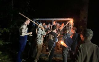 5 Jungs stehen in Dunkelheit mit Fakeln und Stöcken vor einer Gitterbox in der ein Mann (fiktiver Bürgermeister Mai) eingesperrt ist.