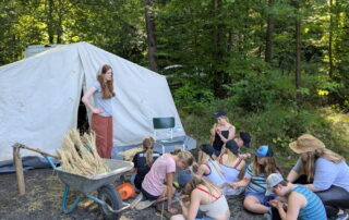 Eine Mädelsgruppe sitzt mit ihren Betreuerinnen vor einem Zelt und bestimmt verschiedene Getreidesorten die in einer Schubkarre aufgereiht zu sehen sind.