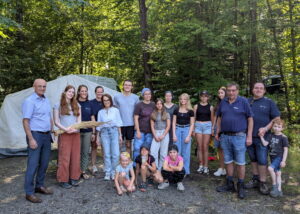 Gruppenfoto von Landrat Wilhelm Schneider, Maya Davey, Familie Göller, Eva Pfeil, Petra Erickson, Wolfgang Winter, Lagerleitung Jonas Erickson und den Zeltbetreuerinnen Elsa Caupert und Doro Mücke gemeinsam mit ihrer Zeltgruppe vor dem Materialzelt am Zeltplatz in Reutersbrunn.
