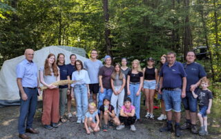Gruppenfoto von Landrat Wilhelm Schneider, Maya Davey, Familie Göller, Eva Pfeil, Petra Erickson, Wolfgang Winter, Lagerleitung Jonas Erickson und den Zeltbetreuerinnen Elsa Caupert und Doro Mücke gemeinsam mit ihrer Zeltgruppe vor dem Materialzelt am Zeltplatz in Reutersbrunn.