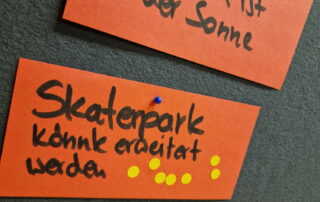 Auf eine grauen Pinnwand sind 2 Moderationskarten gepinnt "Pausenhof ist in der Sonne" und "Skaterpark könnte erweitert werden". Die Skaterpark-Karte hat viele gelbe Votingpunkte.