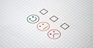 Drei Smileys mit unterschiedlichen Gesichtsausdrücken stehen untereinander mit einem Kästchen zur Abstimmung auf einem karrierten Blatt Papier.
