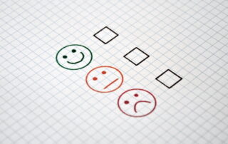 Drei Smileys mit unterschiedlichen Gesichtsausdrücken stehen untereinander mit einem Kästchen zur Abstimmung auf einem karrierten Blatt Papier.