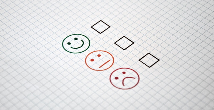 Drei Smileys mit unterschiedlichen Gesichtsausdrücken stehen untereinander mit einem Kästchen zur Abstimmung auf einem karrierten Blatt Papier.