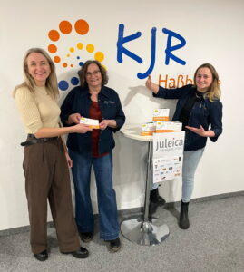 Elvira Jungmann (KJR Verwaltungsmitarbeiterin), Susanne Makowski (KJR Vorsitzende) und Anna-Maria Gebhardt (KJR Vorstandsmitglied, Ressort Ehrenamt) stehen an einem Stehtisch und stellen das neue JULEICA-Gutscheinheft 2026 für den Landkreis Haßberge vor.